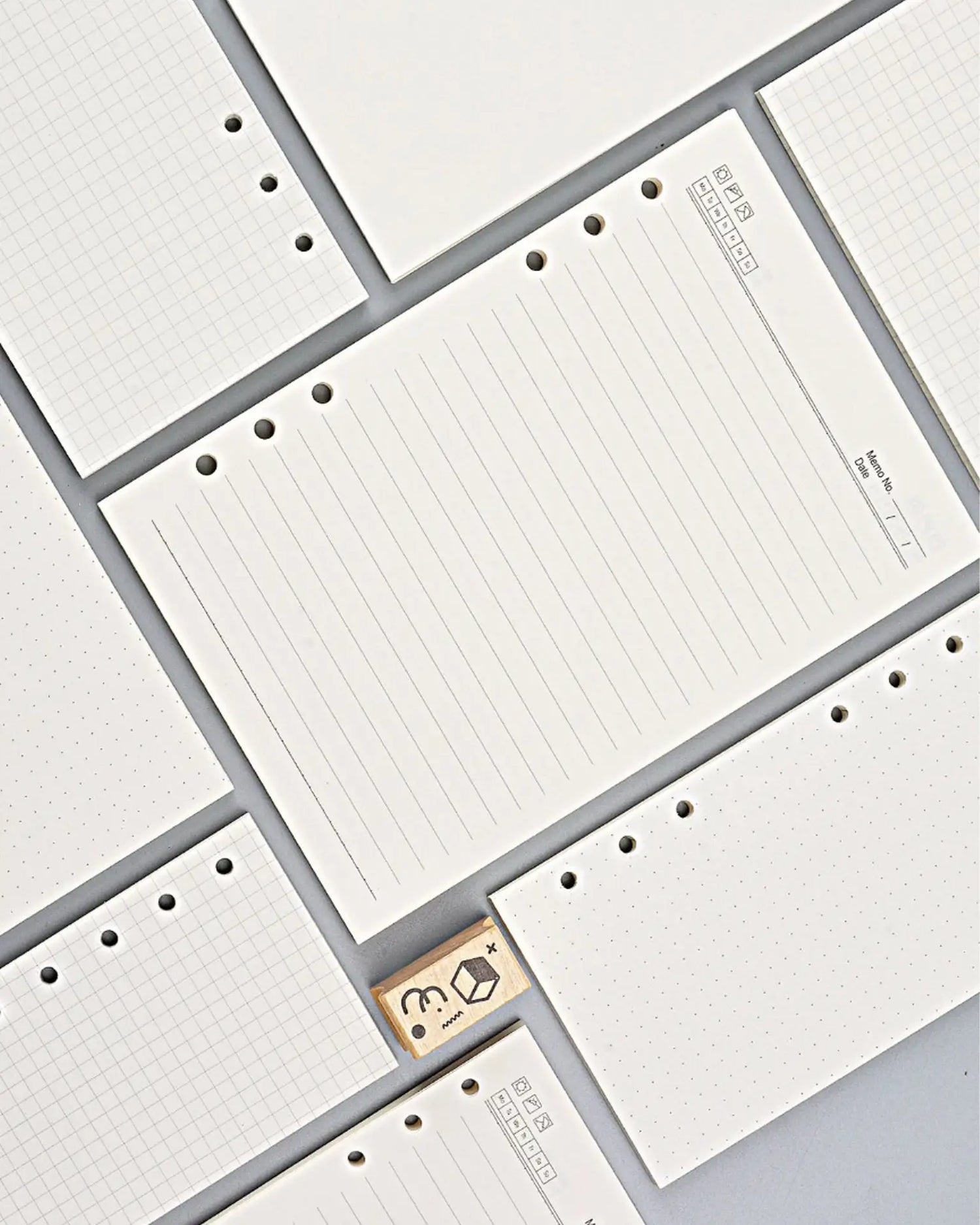 White lined and graph-grid planner inserts arranged in a flat-lay pattern on a light gray background, with one small wooden stamp visible.