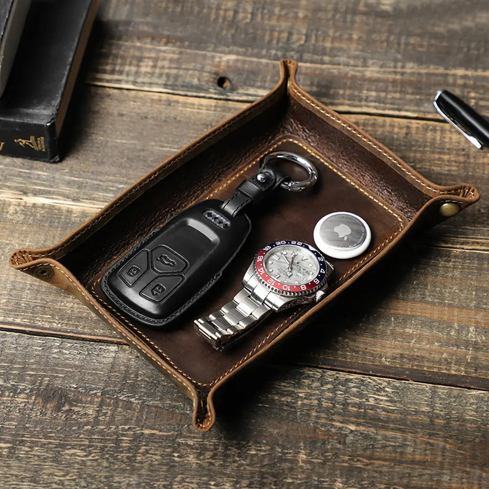 Brown leather tray with key fob, watch, and coins on a wooden surface