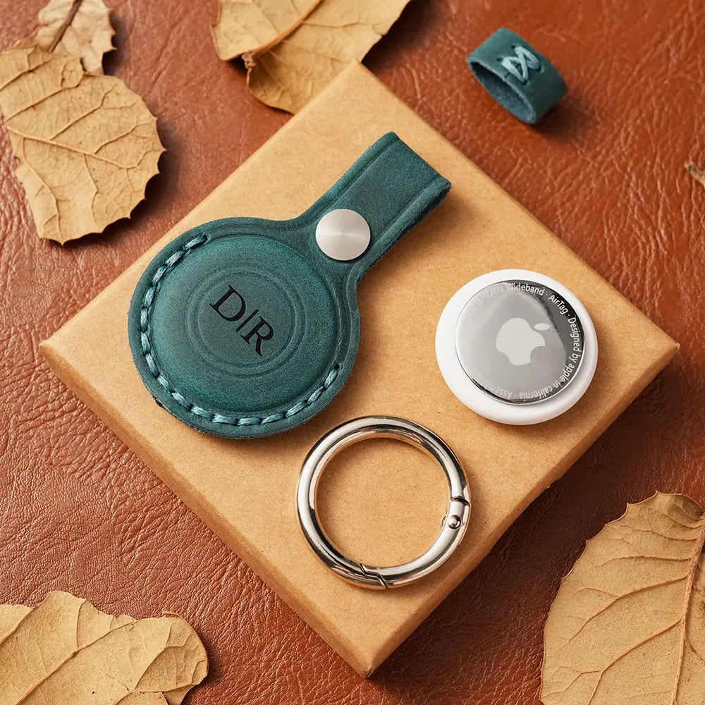 Green leather case with 'DIR' branding and silver ringon a brown AirTag case with leaves.