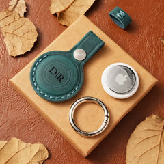 Green leather case with 'DIR' branding and silver ringon a brown AirTag case with leaves.