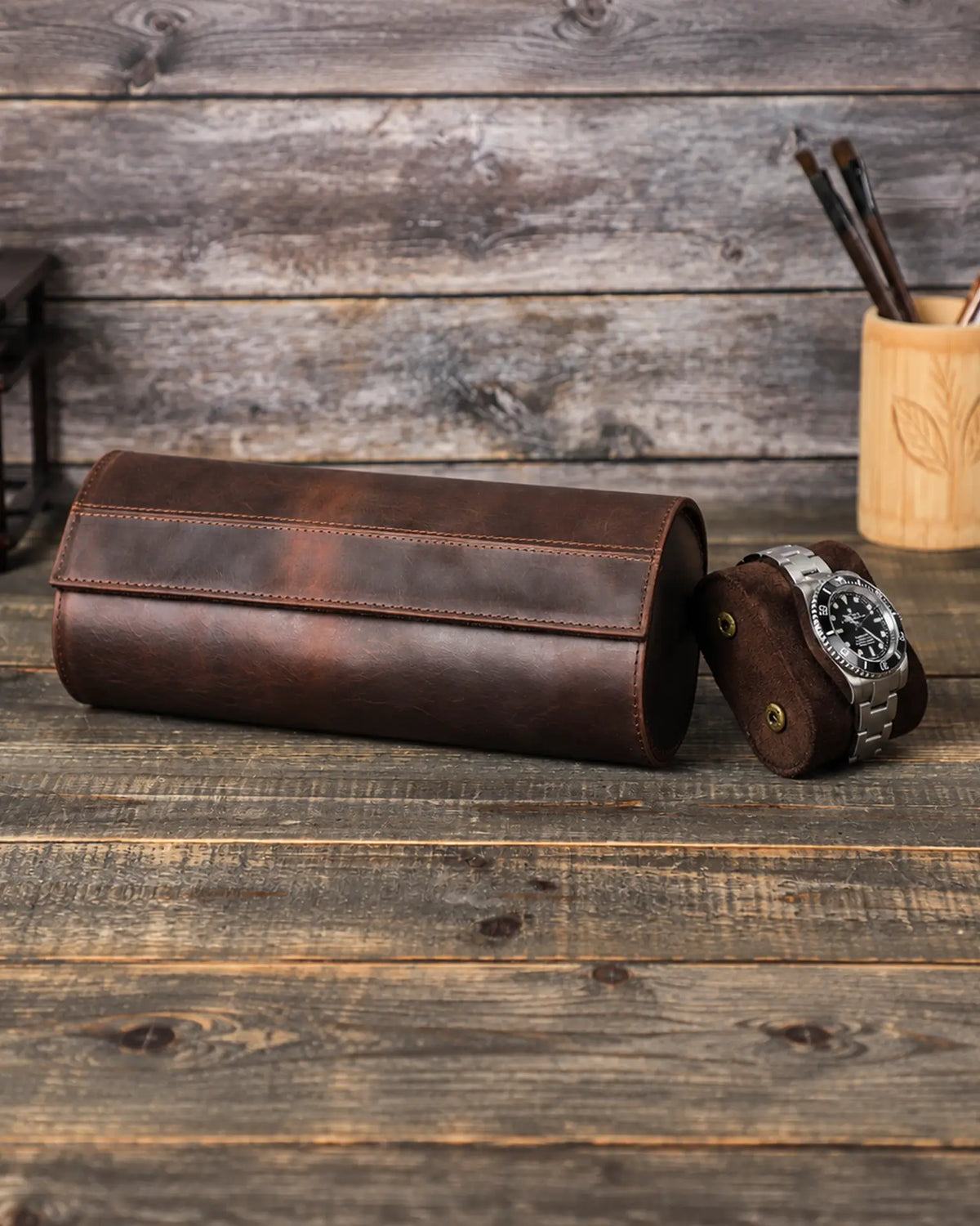 Closed brown triple watch roll beside a removable cushion holding a metal-bracelet watch.