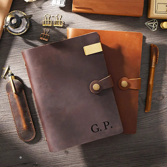 Brown leather notebook with gold accents and 'G.P.' initials on a wooden surface with office supplies.