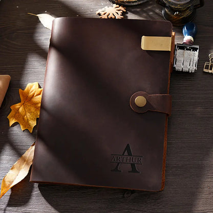 A brown leather notebook with an engraved "Arthur" logo is placed on a wooden surface, surrounded by leaves and stationery items.