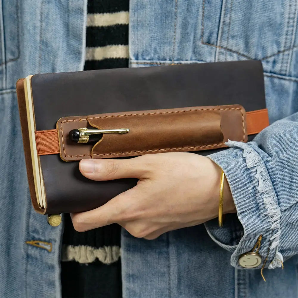 Hand holding a brown leather notebook with a pen holder against a denim background